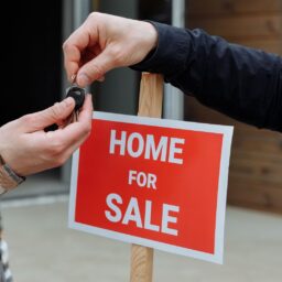 Close-up of a home for sale sign with people exchanging keys.